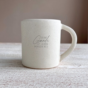 Könnte beinhalten: Weiße Keramiktasse mit gesprenkeltem Finish. Die Tasse hat eine schwarze Aufschrift mit dem Text "Seine Gnade ist jeden Morgen neu".