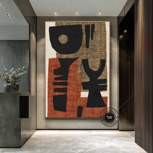 May include: Large framed abstract art piece with a beige, rust, and black color palette. The artwork features geometric shapes and textures, creating a modern and artistic aesthetic. The piece is displayed in a well-lit hallway.
