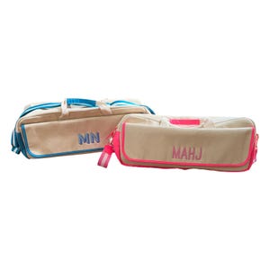 Op de afbeelding: Twee beige canvas duffeltassen. Eén heeft blauwe randen en handvatten, met de initialen "MN" in blauw geborduurd. De andere heeft roze randen en handvatten, met de initialen "MAHJ" in roze geborduurd. Beide tassen hebben een rechthoekige vorm.