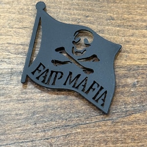May include: Black flag-shaped emblem with a skull and crossbones design. The words "FAIR MAFIA" are cut out at the bottom of the flag. The emblem is set against a wooden background.