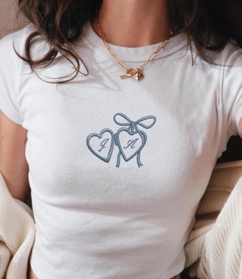 May include: White t-shirt with a crew neck and short sleeves. The shirt features an embroidered design of two blue hearts, each with a letter, connected by a bow. A gold necklace with a pendant is also visible.
