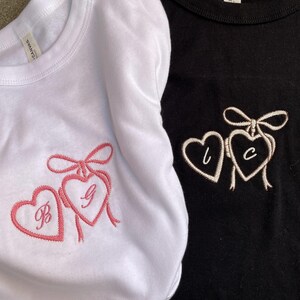 May include: Two t-shirts, one white and one black, each featuring embroidered hearts. The white shirt has pink embroidery with the letters "B" and "G". The black shirt has white embroidery with the letters "L" and "C".