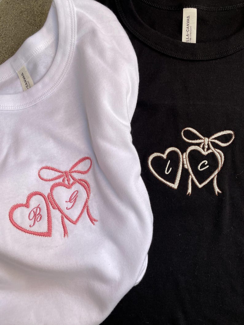 May include: Two t-shirts, one white and one black, each featuring embroidered hearts. The white shirt has pink embroidery with the letters "B" and "G". The black shirt has white embroidery with the letters "L" and "C".