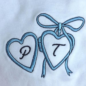 May include: White fabric with two light blue embroidered hearts connected by a bow. Each heart contains a black letter, "P" and "T". The embroidery has a textured appearance.