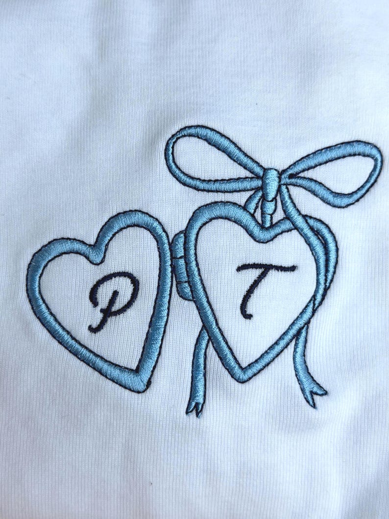 May include: White fabric with two light blue embroidered hearts connected by a bow. Each heart contains a black letter, "P" and "T". The embroidery has a textured appearance.