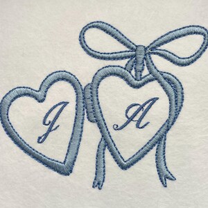 May include: Embroidered design featuring two connected blue hearts with the letters "J" and "A" inside. A blue bow and ribbons adorn the top. The design is on a white background.