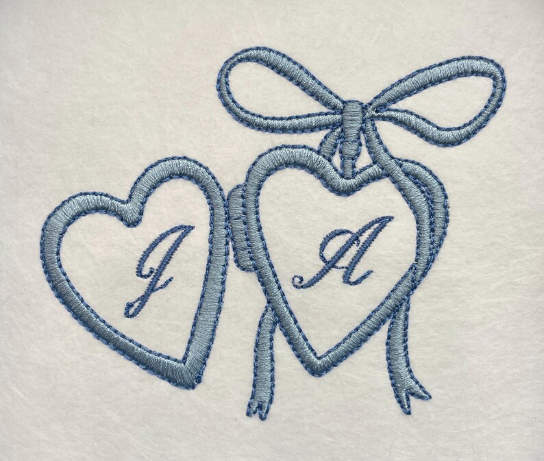 May include: Embroidered design featuring two connected blue hearts with the letters "J" and "A" inside. A blue bow and ribbons adorn the top. The design is on a white background.