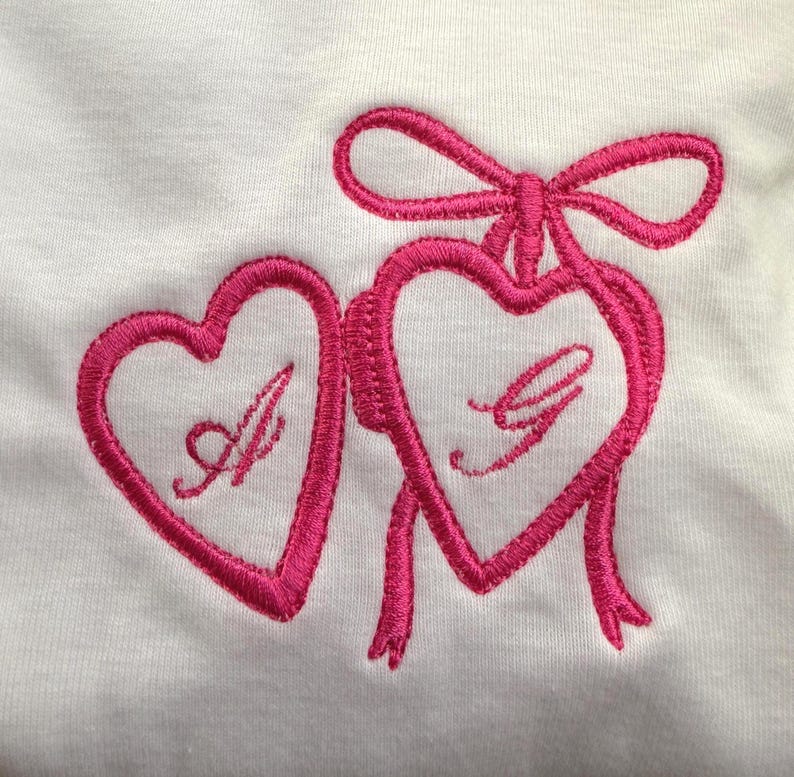 May include: White fabric with a pink embroidered design featuring two hearts connected by a ribbon and bow. The left heart has the letter "A" and the right heart has the letter "G".