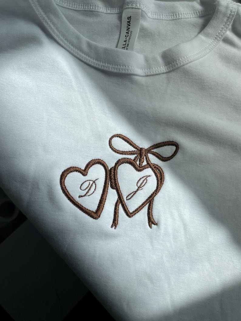 May include: White t-shirt with a brown embroidered design of two connected hearts, each with a letter inside. The hearts are tied together with a bow and ribbons. The tag reads "BELLA+CANVAS."