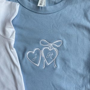 May include: Light blue t-shirt with a white embroidered design of two hearts tied together with a bow. The heart on the left has the letter "J" and the heart on the right has the letter "H". A white t-shirt is partially visible on the left.