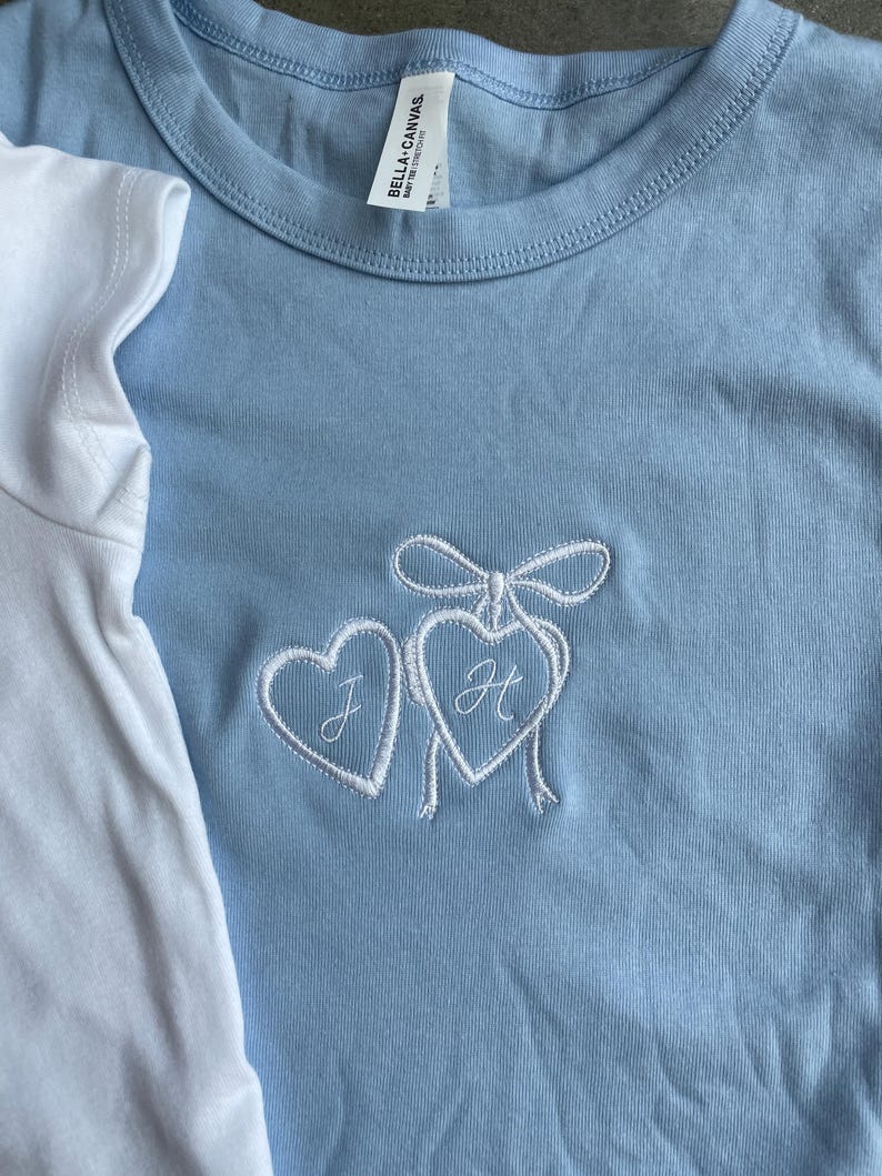 May include: Light blue t-shirt with a white embroidered design of two hearts tied together with a bow. The heart on the left has the letter "J" and the heart on the right has the letter "H". A white t-shirt is partially visible on the left.