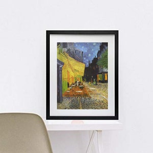 May include: A framed print of Van Gogh's "Cafe Terrace at Night" in a black frame. The artwork depicts a nighttime street scene with a yellow cafe, tables, and figures. The print is displayed on a white desk with a modern chair.