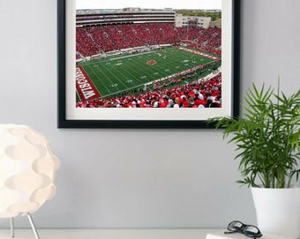 Può includere: Stampa incorniciata di uno stadio di football pieno di tifosi vestiti di rosso. Il campo verde ha linee bianche e la parola "WISCONSIN" in rosso. Le tribune sono piene, creando una scena vivace.