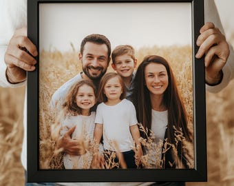 Può includere: Un ritratto di famiglia incorniciato con una famiglia sorridente di cinque persone in un campo di grano. La cornice nera circonda la fotografia, che cattura le espressioni gioiose della famiglia. L'immagine è tenuta da una persona che indossa un maglione bianco e jeans blu.