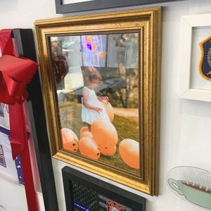 May include: A framed photograph in a gold ornate frame. The image shows a young child in a white dress with orange balloons. The frame is mounted on a white wall, alongside other framed items and decorative elements.