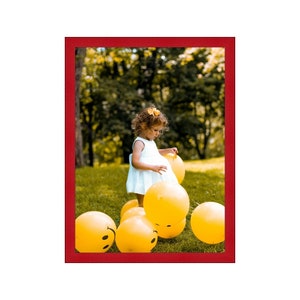 May include: A young girl in a white dress plays with yellow balloons in a grassy park. The balloons have smiley faces on them.