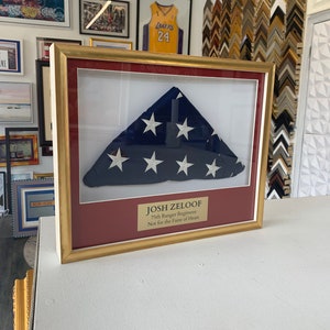 Könnte beinhalten: Eine gerahmte amerikanische Flagge mit einem roten Passepartout und einem goldenen Rahmen. Die Flagge ist zu einem Dreieck gefaltet und hat weiße Sterne auf einem blauen Feld. Eine Plakette unter der Flagge lautet "JOSH ZELOOF 75th Ranger Regiment Not for the Faint of Heart."