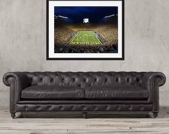 Può includere: Fotografia incorniciata di uno stadio di football di notte, con la parola "MICHIGAN" visibile sul campo. L'immagine è ambientata contro un muro grigio, sopra un divano in pelle marrone scuro con bottoni.