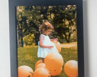 May include: A young girl in a white dress stands on a pile of yellow smiley face balloons in a grassy field.