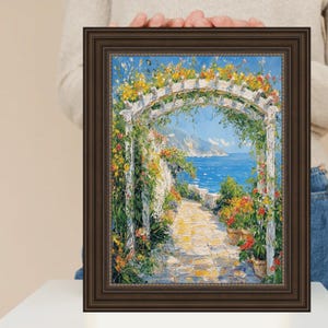 May include: A framed oil painting shows a coastal scene with a white pergola covered in yellow flowers. A stone path leads to the sea, with potted flowers on either side. The frame is a dark brown colour.