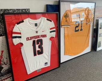 Può includere: Due maglie da football americano incorniciate. Una è bianca con dettagli rossi e neri, con la scritta "CLIFFSIDE PARK" e il numero "13". L'altra è arancione con il numero "21". Entrambe in cornici nere.