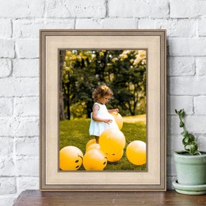 May include: A young girl in a white dress plays with yellow balloons in a grassy park. The balloons have smiley faces on them.