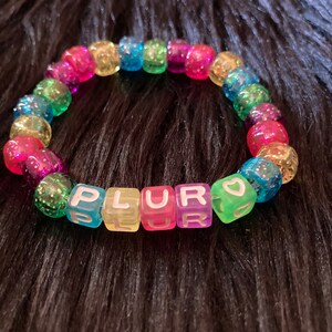 Könnte beinhalten: Ein Regenbogen-Perlenarmband mit dem Wort "PLUR" in bunten Buchstabenperlen. Das Armband enthält auch eine kleine Herzperle.