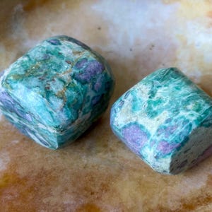 May include: Two polished, square-cut gemstones. The stones display a mix of teal, green, and purple hues, with a marbled appearance. They rest on a light brown, textured surface, creating a natural and earthy aesthetic.