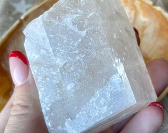 Może przedstawiać: Półprzezroczysty, biały okaz minerału w kształcie sześcianu o krystalicznej, teksturowanej powierzchni. Kamień posiada naturalne płaszczyzny łupliwości i wewnętrzne pęknięcia, wykazując półprzezroczysty wygląd o różnym stopniu przejrzystości.