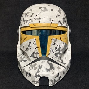 May include: A white and gray camouflage helmet with yellow accents and black stitching. The helmet has a blue visor.