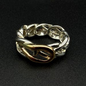 May include: A silver-toned ring with a chain-link design and a gold-colored clasp. The ring is set against a black background, highlighting its metallic sheen and intricate details. The ring's design is a blend of textures and colors.