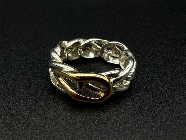 May include: A silver-toned ring with a chain-link design and a gold-colored clasp. The ring is set against a black background, highlighting its metallic sheen and intricate details. The ring's design is a blend of textures and colors.