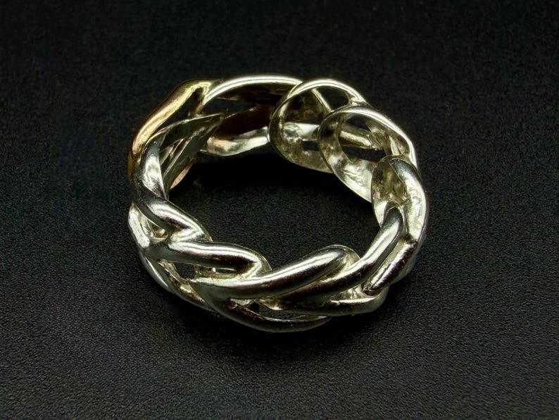 May include: A silver-toned chain link ring. The ring features a continuous loop of interlocking chain links, with a gold-colored section visible. The ring is set against a dark background.