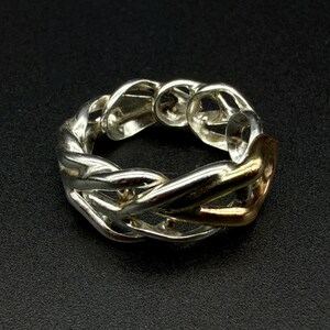 May include: A silver-toned ring with a braided design, featuring a gold-colored accent. The ring is set against a dark background, highlighting its intricate details and metallic sheen. The ring's design is a continuous loop, showcasing a blend of textures.