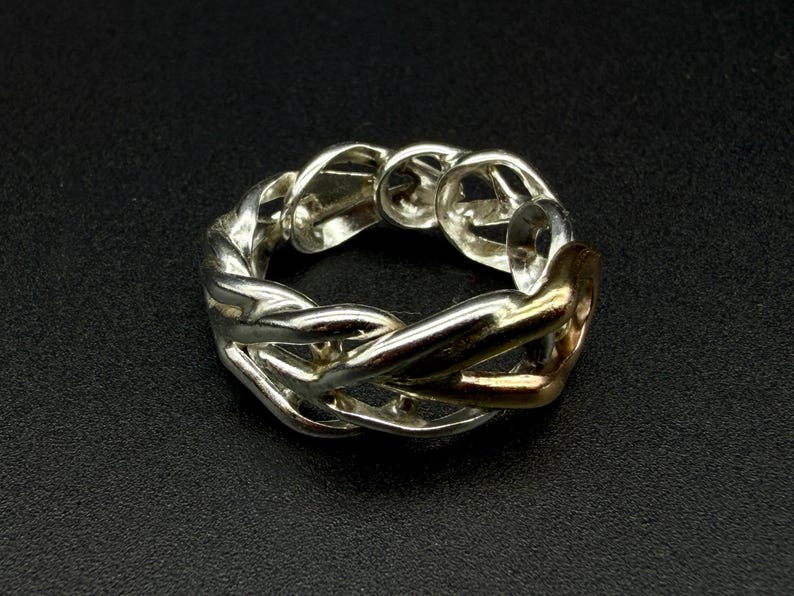 May include: A silver-toned ring with a braided design, featuring a gold-colored accent. The ring is set against a dark background, highlighting its intricate details and metallic sheen. The ring's design is a continuous loop, showcasing a blend of textures.