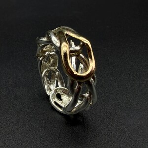 May include: A silver-toned ring with a chain-link design, featuring a gold-toned central link. The ring is set against a dark background, highlighting its metallic sheen and intricate details. A piece of jewelry.