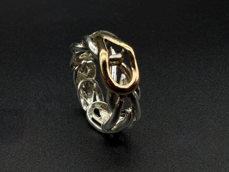 May include: A silver-toned ring with a chain-link design, featuring a gold-toned central link. The ring is set against a dark background, highlighting its metallic sheen and intricate details. A piece of jewelry.