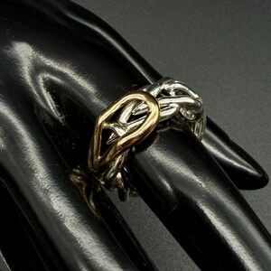 May include: A two-tone ring with a gold-colored center and silver-colored interwoven bands. The ring is worn on a finger, showcasing its intricate design and contrasting metallic hues. The ring's design is a modern take on a classic style.