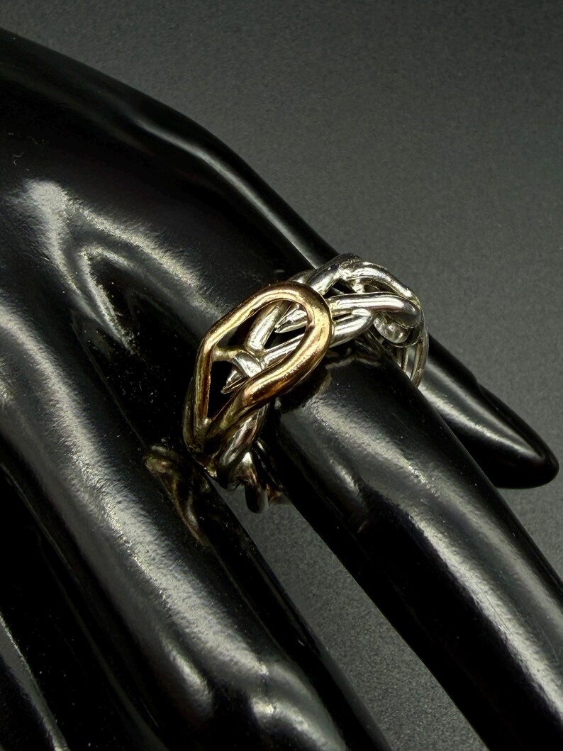 May include: A two-tone ring with a gold-colored center and silver-colored interwoven bands. The ring is worn on a finger, showcasing its intricate design and contrasting metallic hues. The ring's design is a modern take on a classic style.