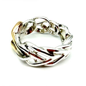 May include: A silver ring with a woven chain design. The ring has a gold-colored section on one side. The ring is set against a white background.