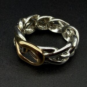 May include: A silver-toned chain link ring with a gold-colored link. The ring is set against a dark background, highlighting its metallic shine. The design features a series of interconnected links, creating a bold and stylish accessory.
