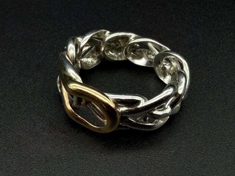 May include: A silver-toned chain link ring with a gold-colored link. The ring is set against a dark background, highlighting its metallic shine. The design features a series of interconnected links, creating a bold and stylish accessory.