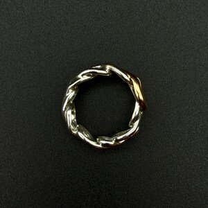 May include: A silver-toned ring with a braided chain design. A section of the ring is gold-toned. The ring is set against a black background.