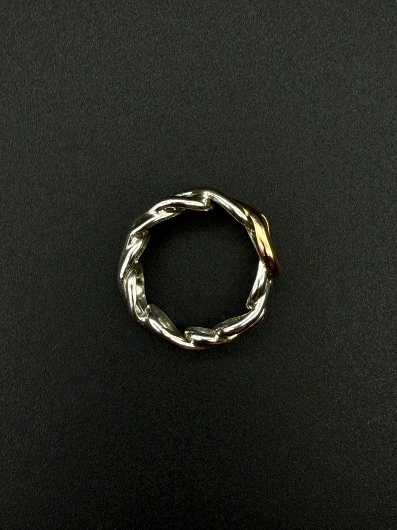 May include: A silver-toned ring with a braided chain design. A section of the ring is gold-toned. The ring is set against a black background.