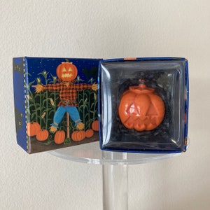 May include: A blue box with a scarecrow and pumpkins on the front. The box contains a small orange pumpkin shaped candle with a carved face.