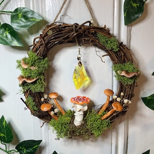 May include: A small, rustic wreath made of twigs and moss, decorated with miniature mushrooms and a crystal pendant. The wreath is hanging on a white door.