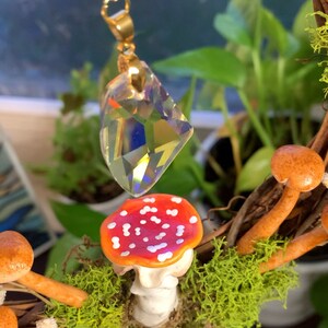 May include: A crystal prism hangs from a gold chain above a red and white mushroom with white spots. The mushroom is surrounded by green moss and brown twigs.