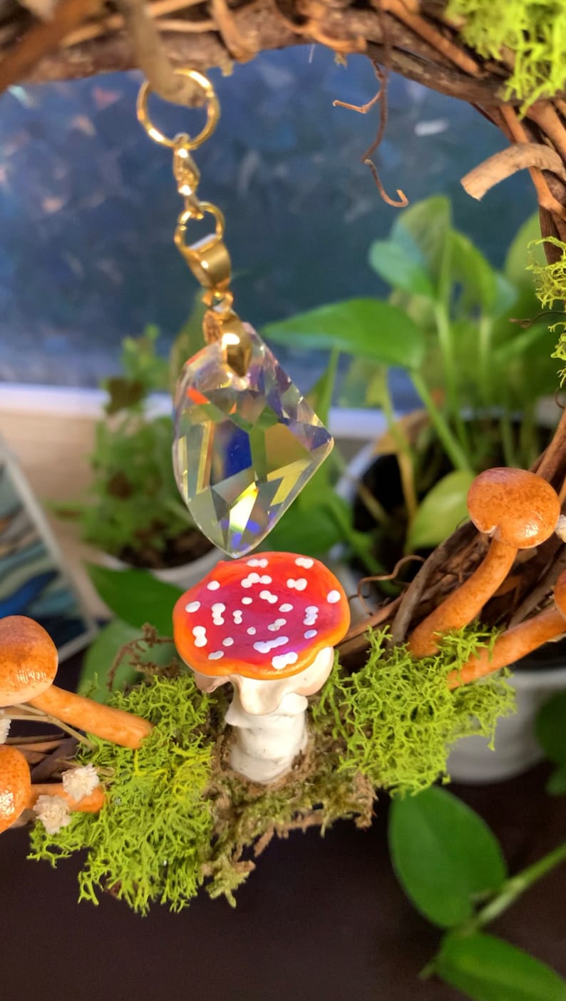 May include: A crystal prism hangs from a gold chain above a red and white mushroom with white spots. The mushroom is surrounded by green moss and brown twigs.