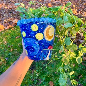 Puede incluir: Una maceta de cerámica azul y amarilla con un diseño de noche estrellada, que incluye una luna creciente y un hongo rojo. La maceta está llena de plantas verdes de hoja.