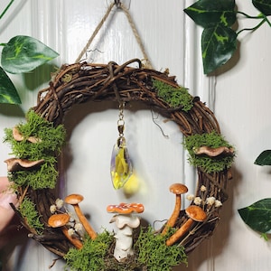 May include: A brown twig wreath with green moss, small white flowers, and brown mushrooms. A yellow crystal hangs from the wreath.